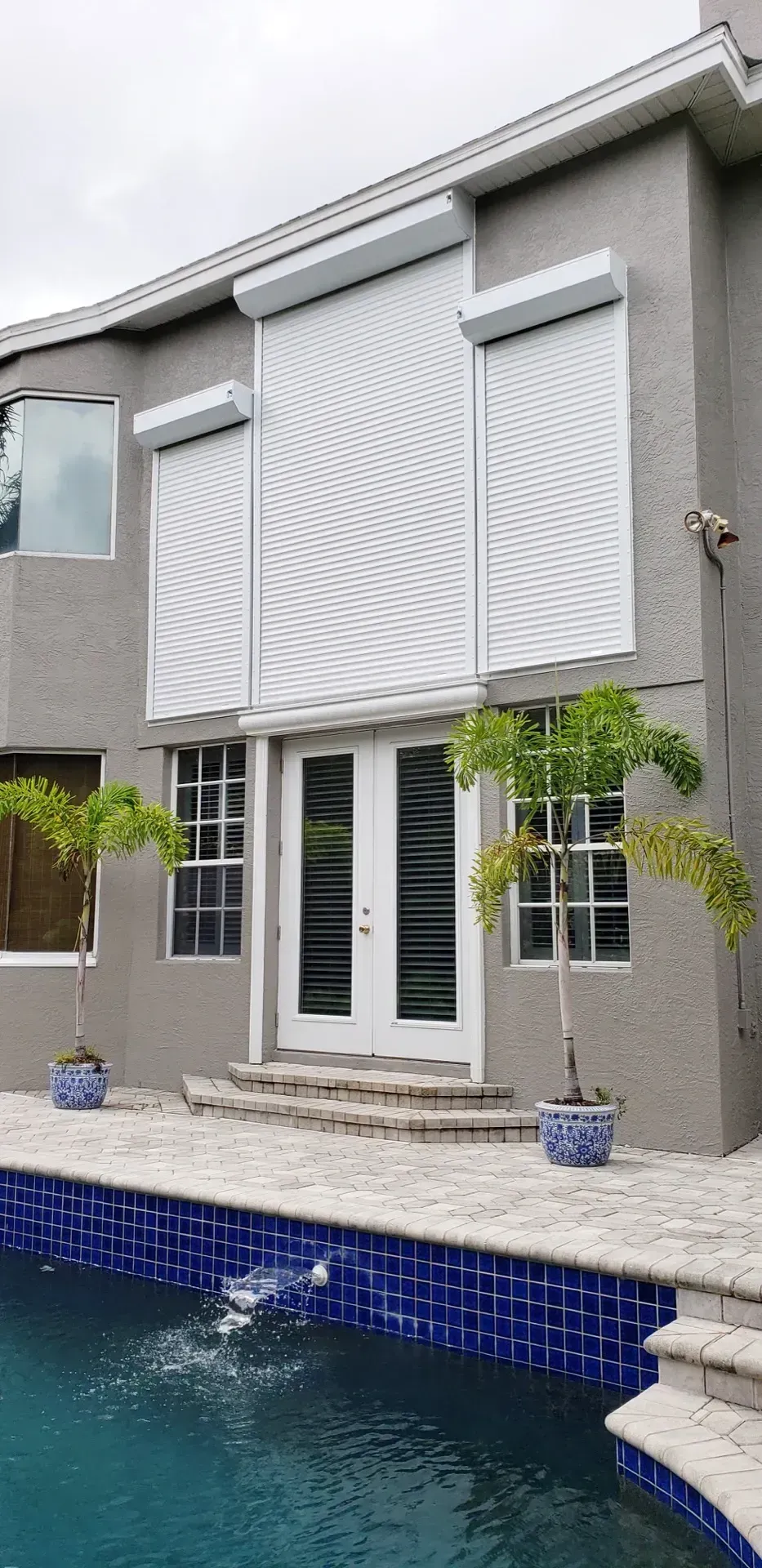 House with pool, hurricane shutters over windows and doors, palm trees in pots.