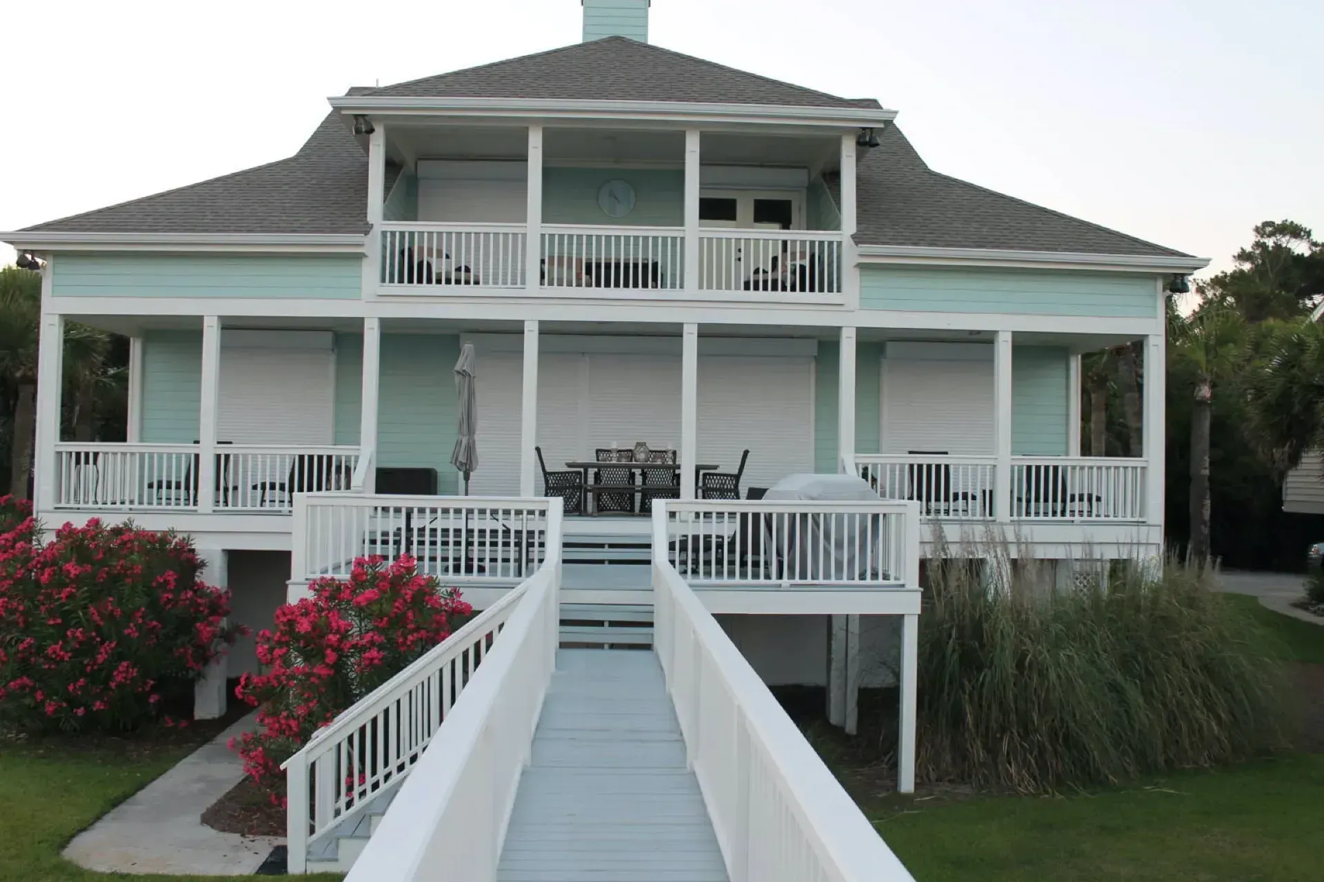 Two-story, light blue coastal house with white railings and closed shutters; walkway leads to the entrance.