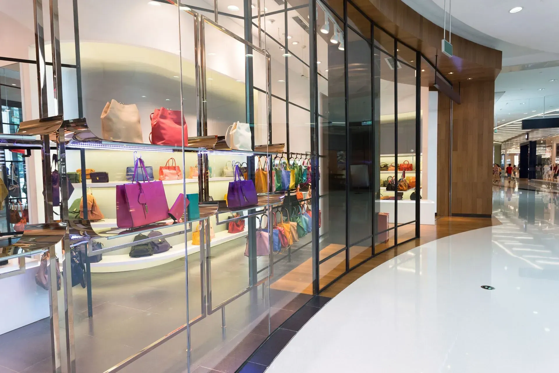 A glass-walled handbag store display with colorful purses and bags in a bright shopping mall.