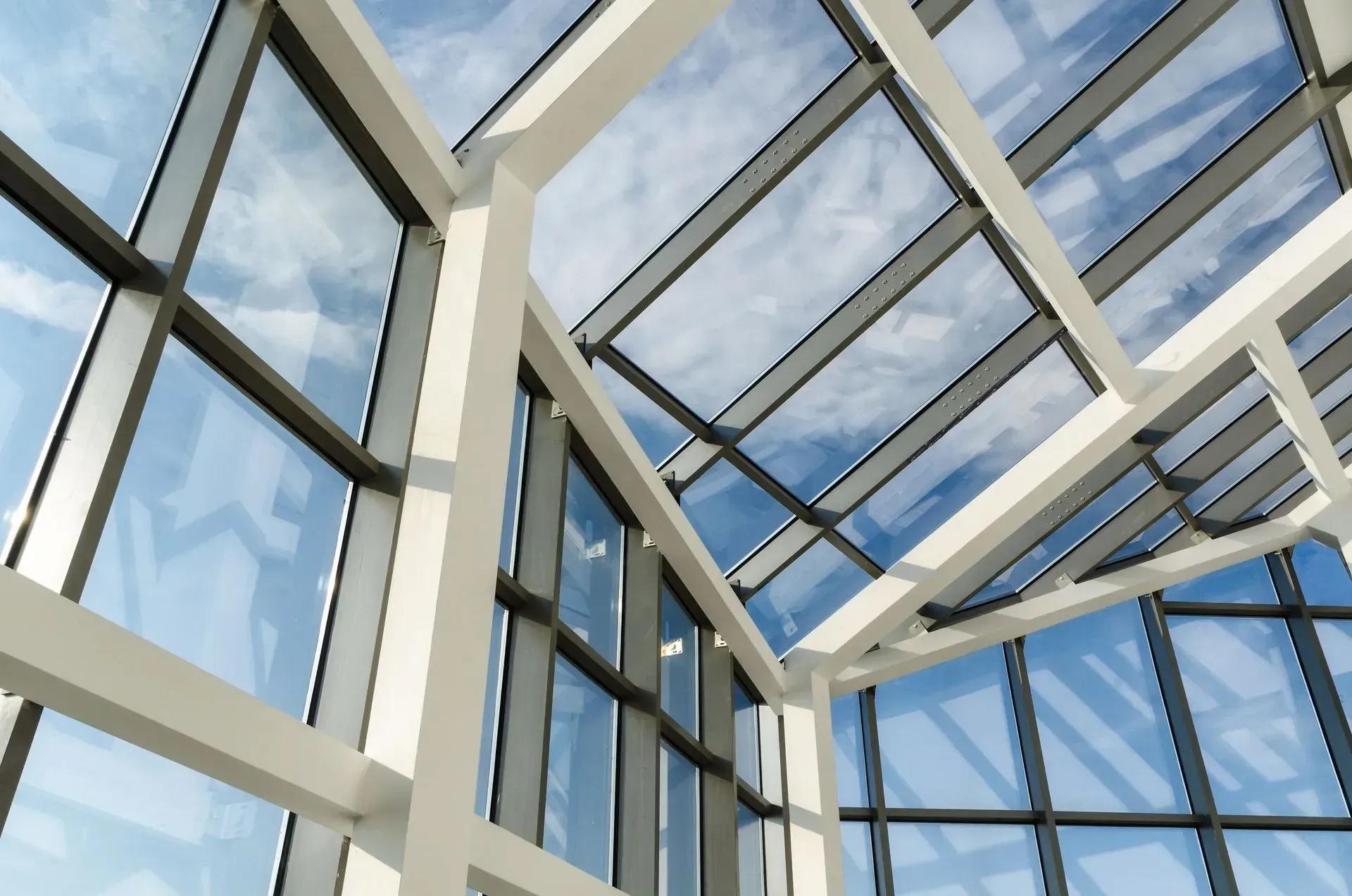 Glass ceiling and wall of a modern building with white support beams reflecting the sky.