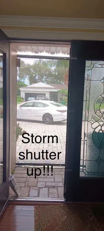 View through a door, storm shutter up. A white car and house are visible through the opening.