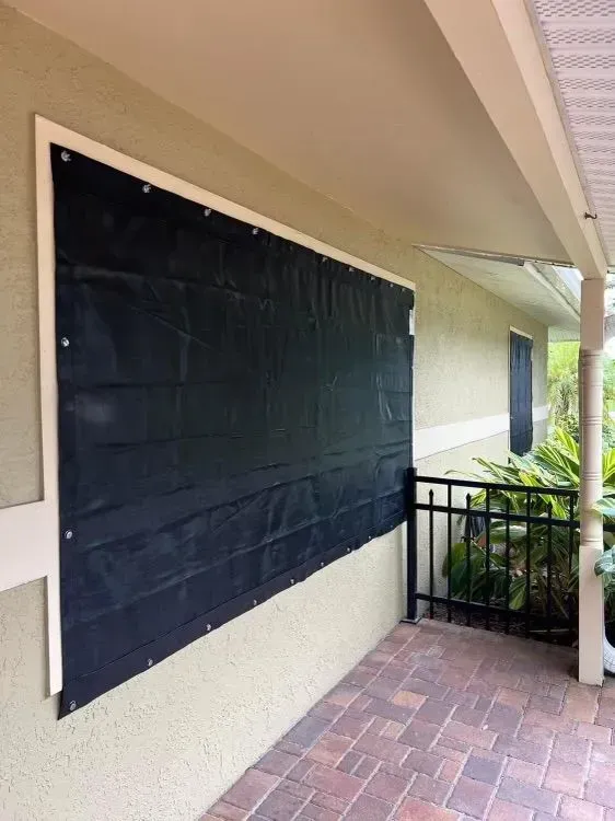 Black storm shutter covering window on beige building. White trim and brick patio visible.