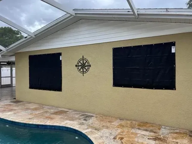 Exterior of a house with two windows covered by black hurricane shutters next to a pool.