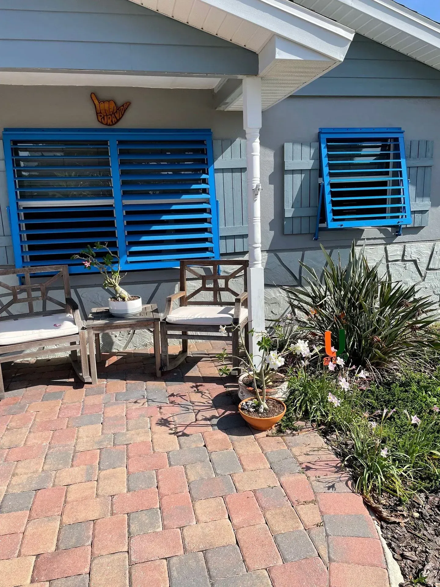 Blue-shuttered house front with porch. Wooden chairs and small table on brick patio. Flowers and greenery.