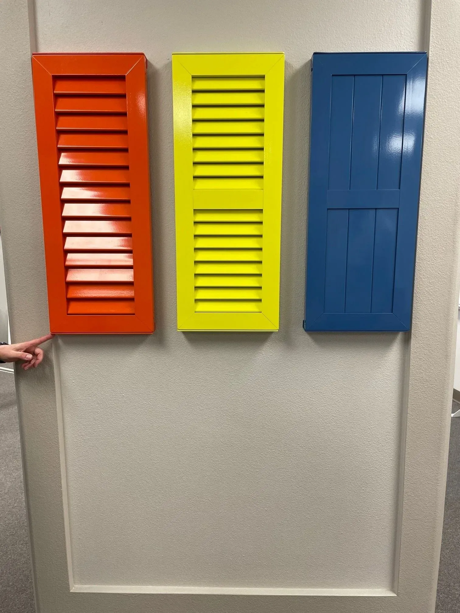 Three brightly colored shutters: orange, yellow, and blue, mounted on a light gray wall.