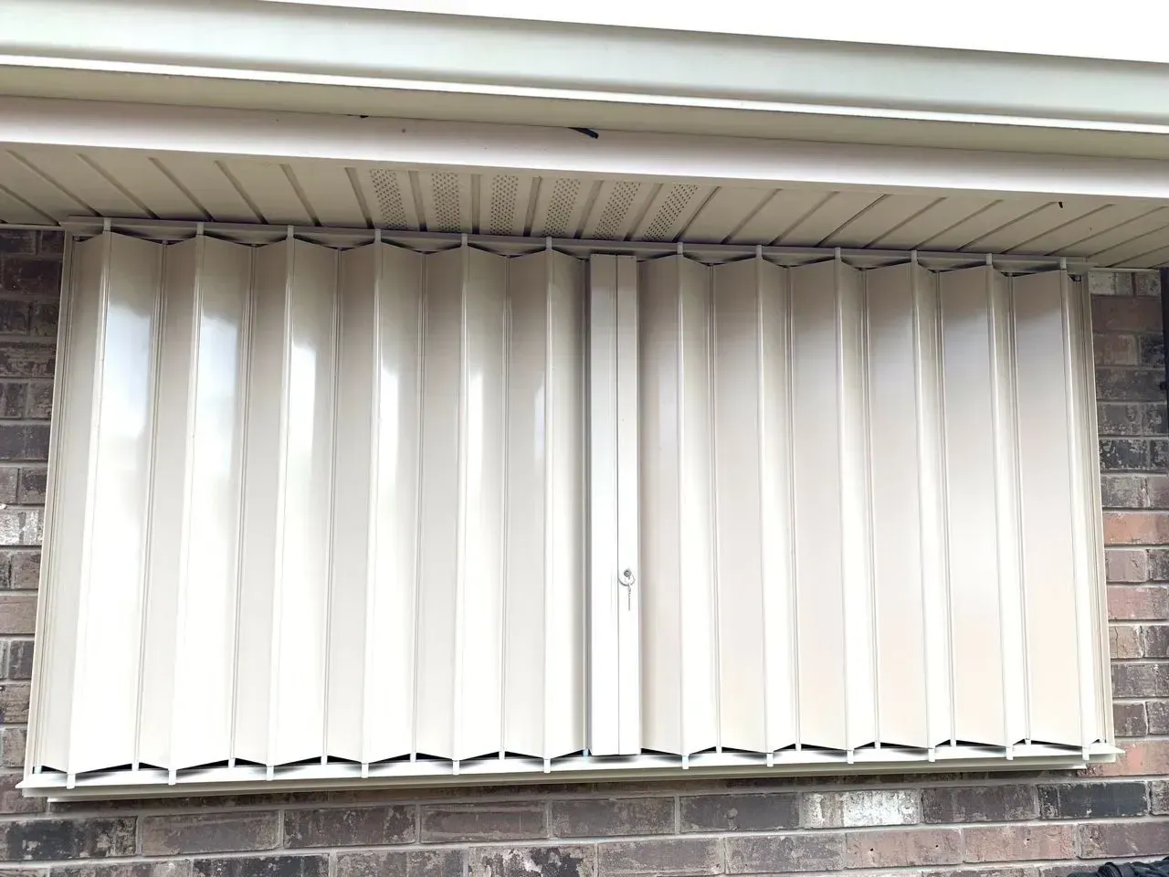 Tan accordion-style storm shutters closed over a window on a brick building, under a white eave.