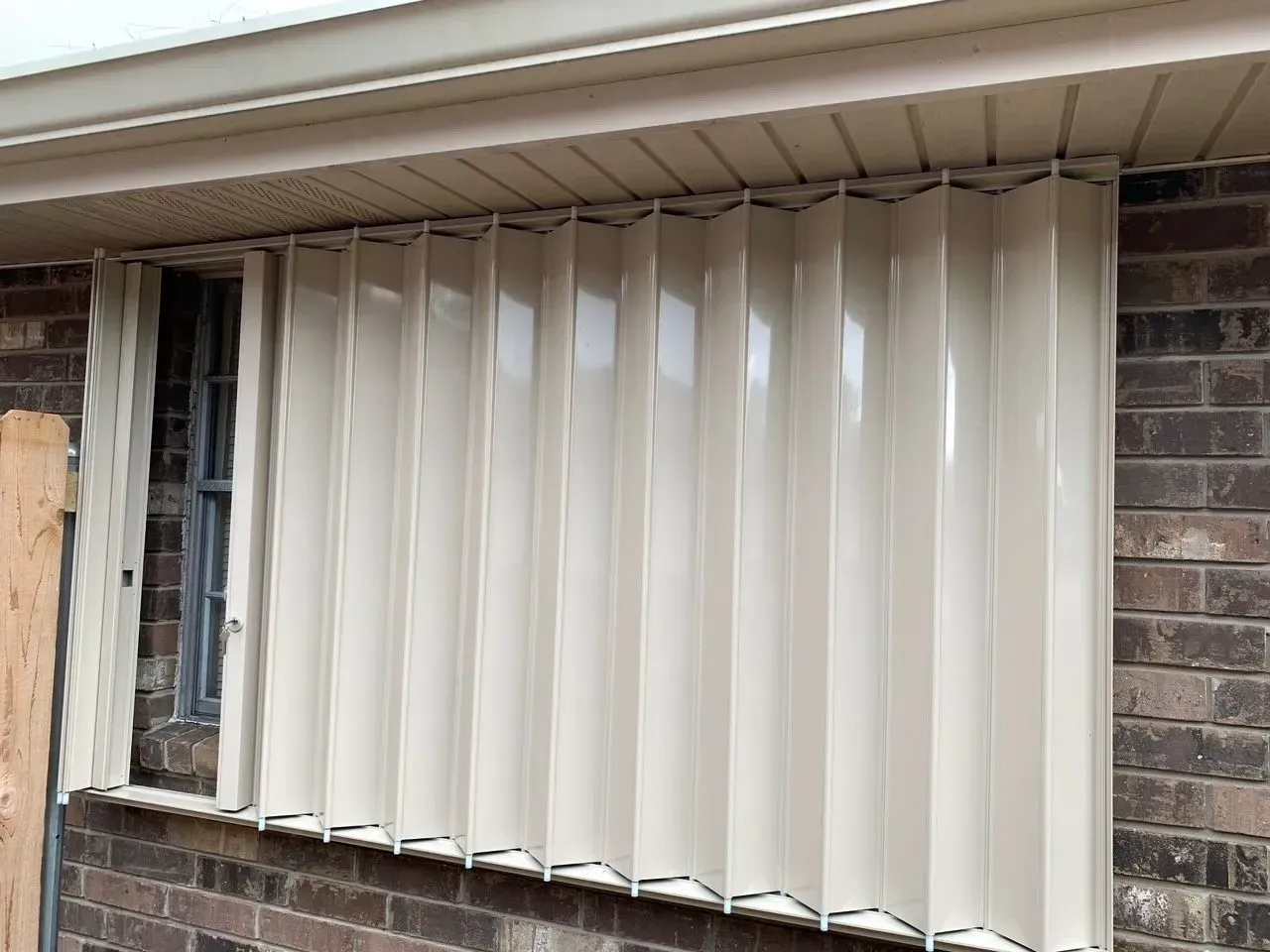 Tan accordion-style hurricane shutters installed over a window on a brick building.