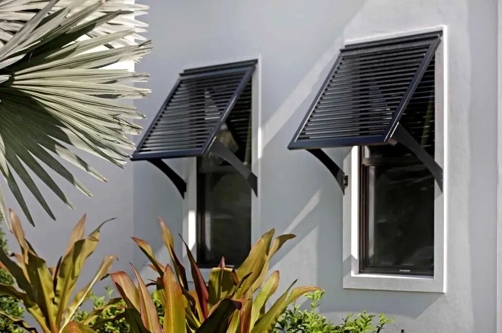 Two windows with open, black, slatted awnings on a white building exterior with greenery.