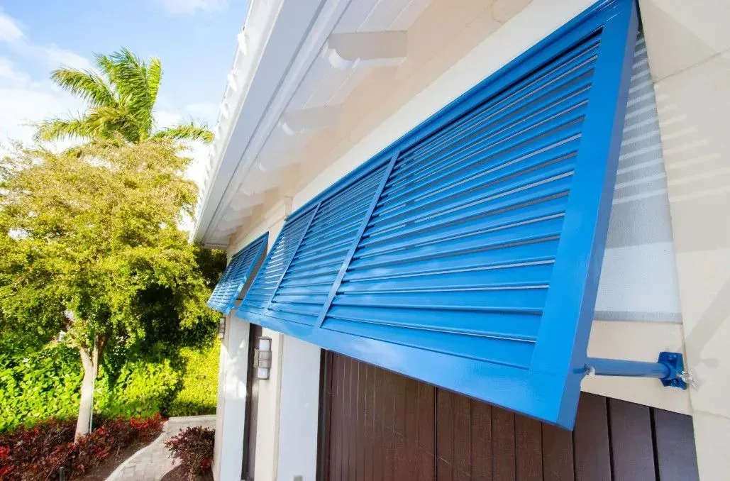 Blue hurricane shutters on a white house, with a tree in the background.