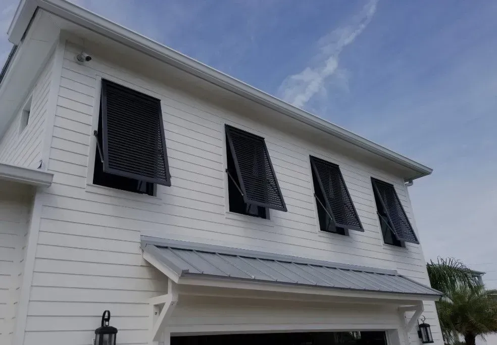 White house with gray roof, black hurricane shutters open on several windows, under a blue sky.