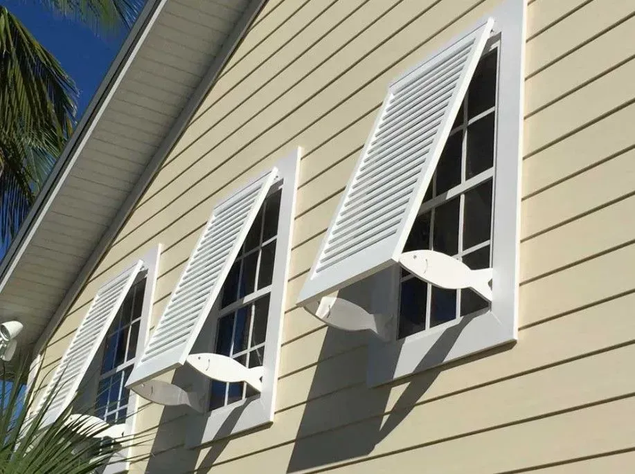 White louvered shutters on a building with triangular windows. Each shutter has a decorative fish shape, set against beige siding.