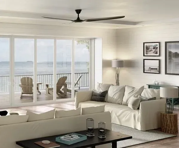 Living room with large windows overlooking a waterfront balcony; white sofas, dark ceiling fan, black framed artwork.