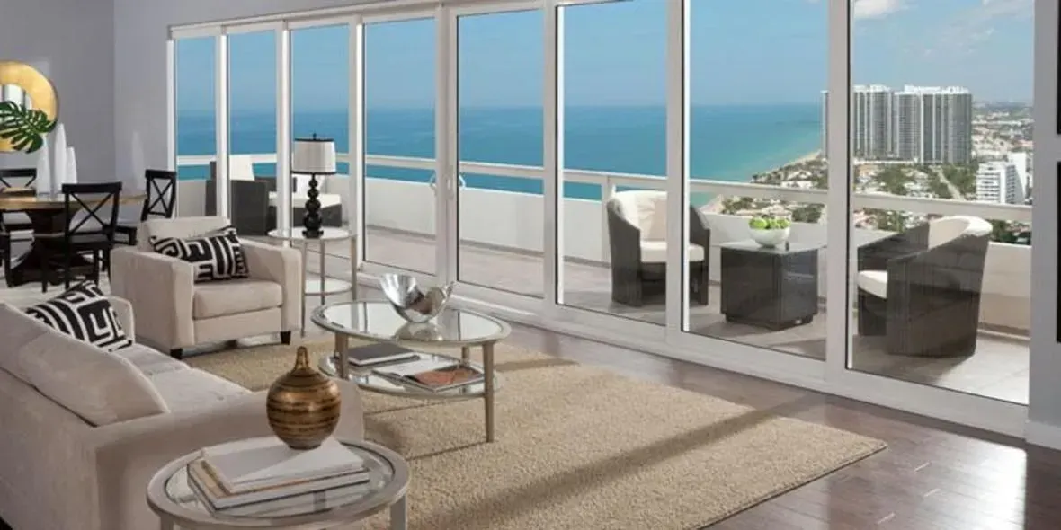 Living room with large windows overlooking a beach and city. Furnished with beige sofa and rug.