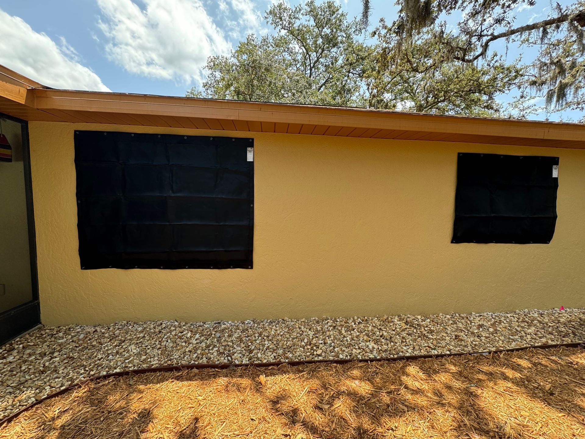 Yellow stucco building with two dark-covered windows, gravel border, and mulch ground cover.