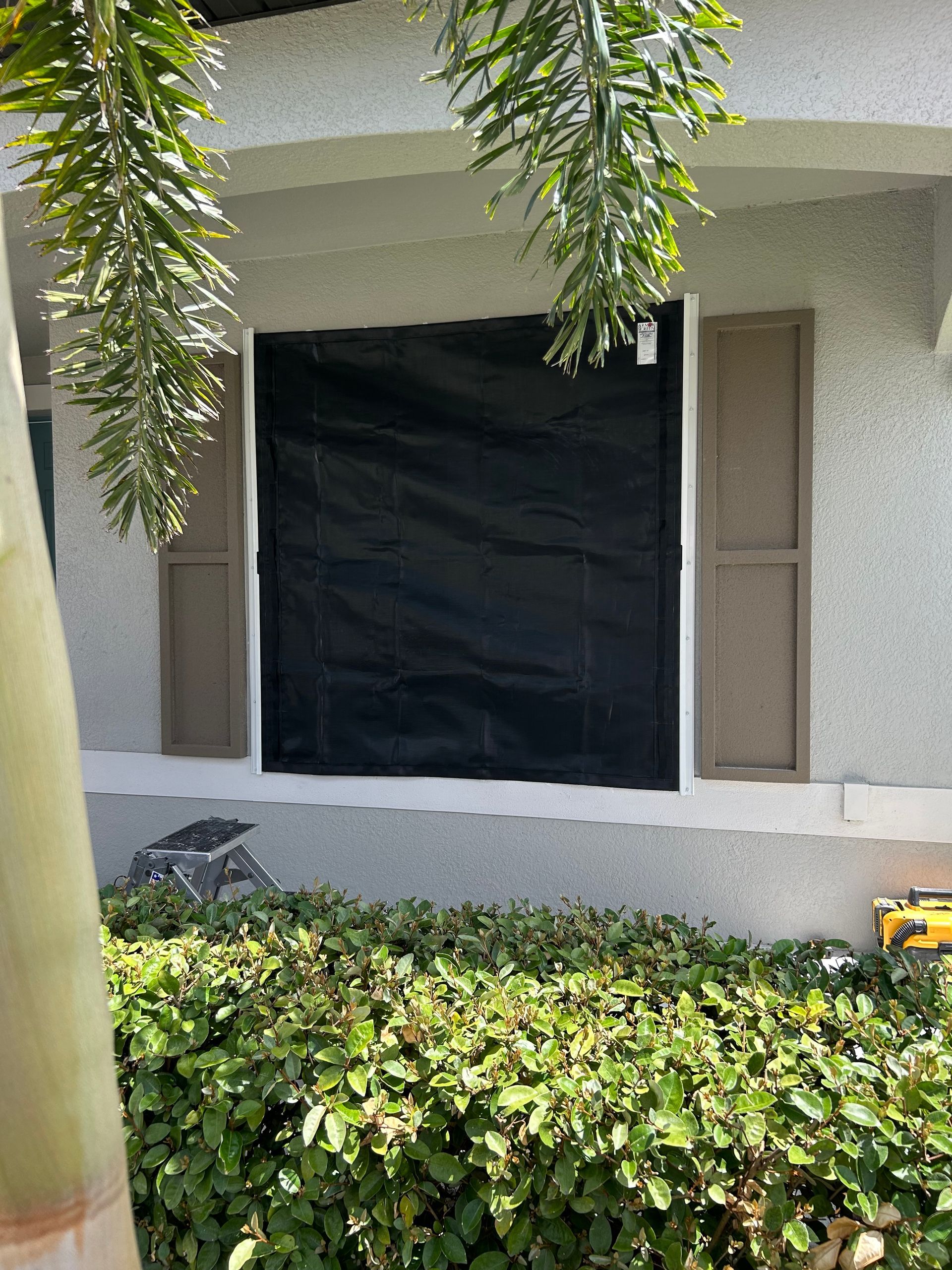 A window covered with a black tarp, flanked by tan shutters, on a stucco building.