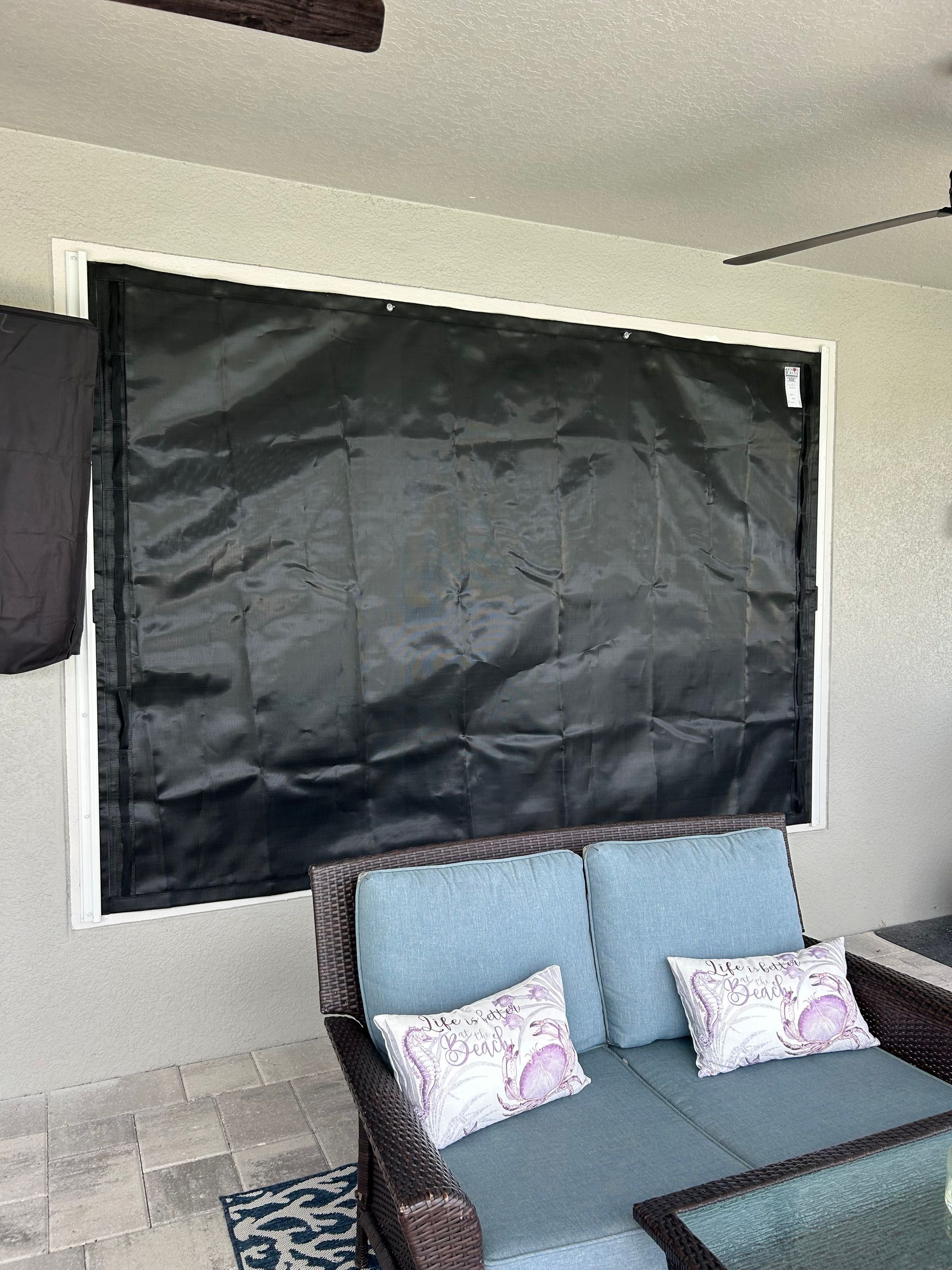 Black storm shutter covering a window on an outdoor patio, above a blue loveseat with pillows.