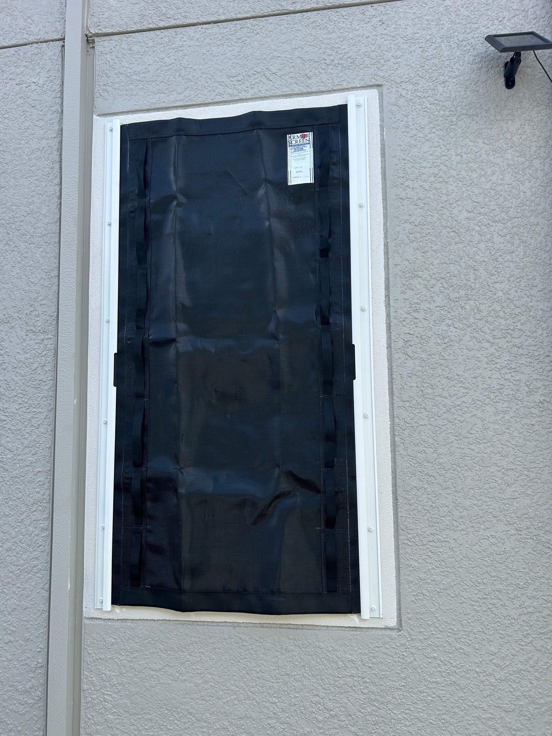 Black covering over a window, secured to a white frame on a textured gray building.
