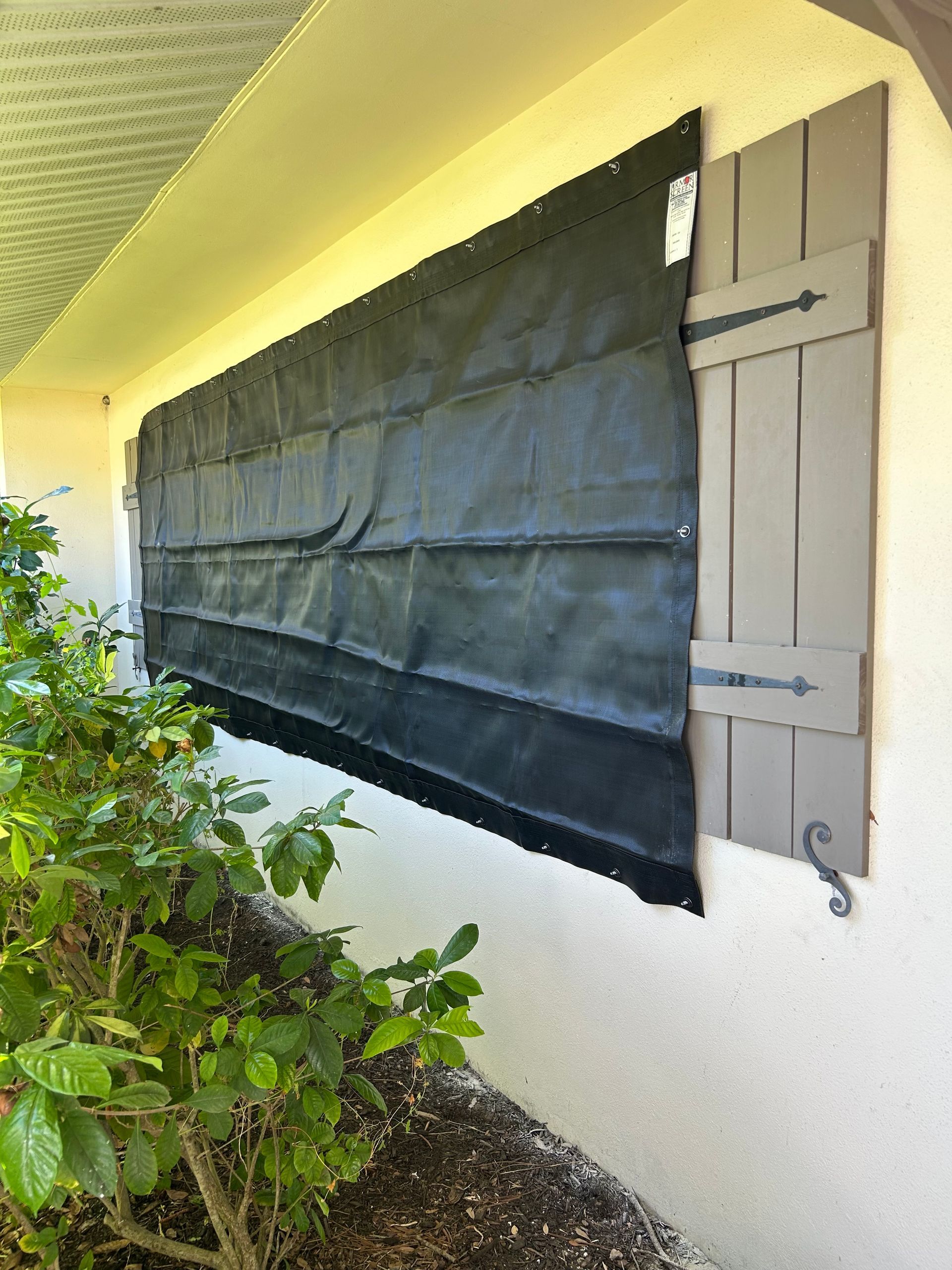 Black storm shutter installed over a window, next to closed gray shutters on a white building.