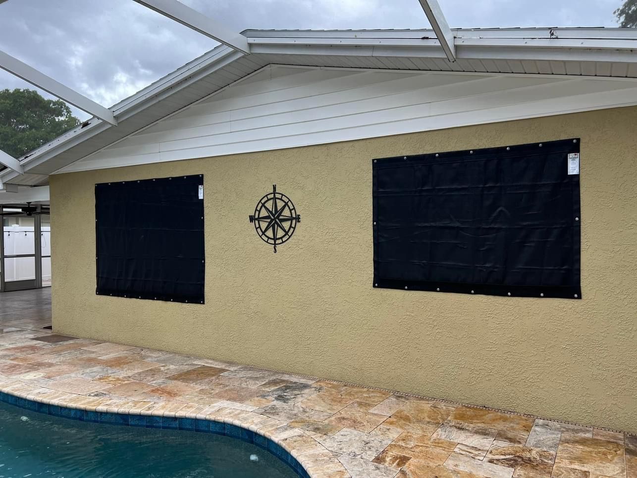 Exterior of a house with two windows covered by black storm shutters; pool in foreground, cloudy sky.