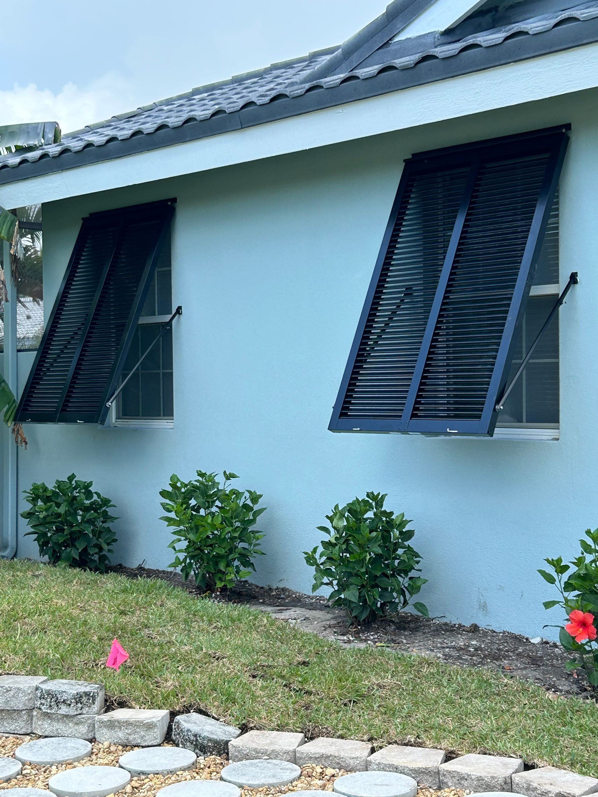 Two windows with black louvered shutters on a light blue building, set on a grassy lawn.