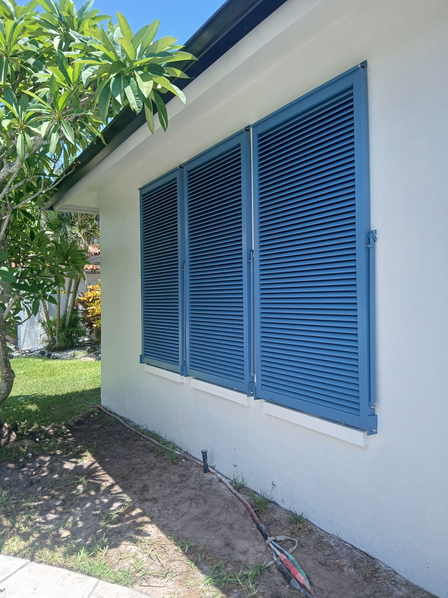 Blue louvered shutters on white house exterior. Green grass and tree in the background.