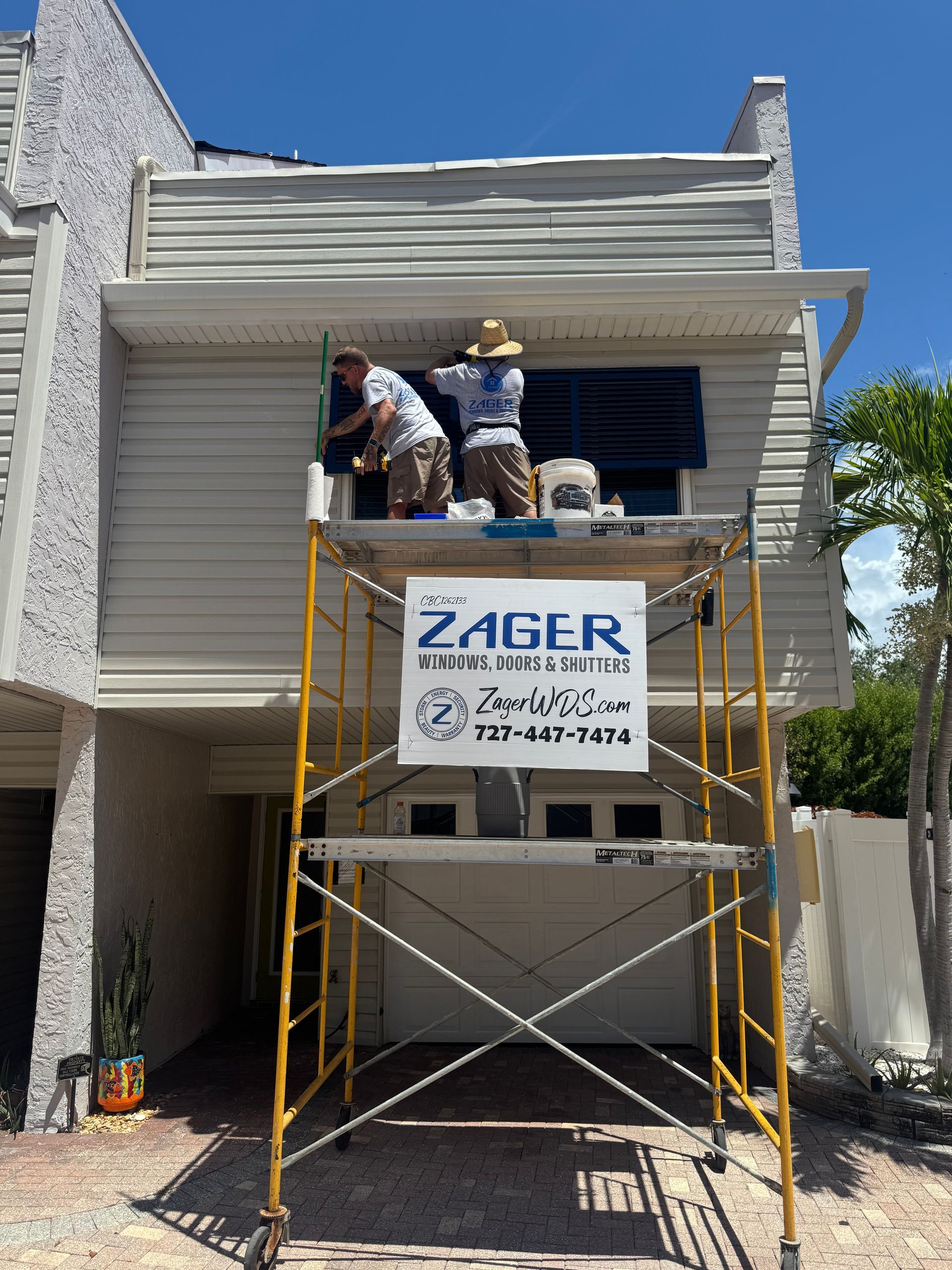 Two workers install hurricane shutters on a building's window, using a scaffolding in front of a garage.