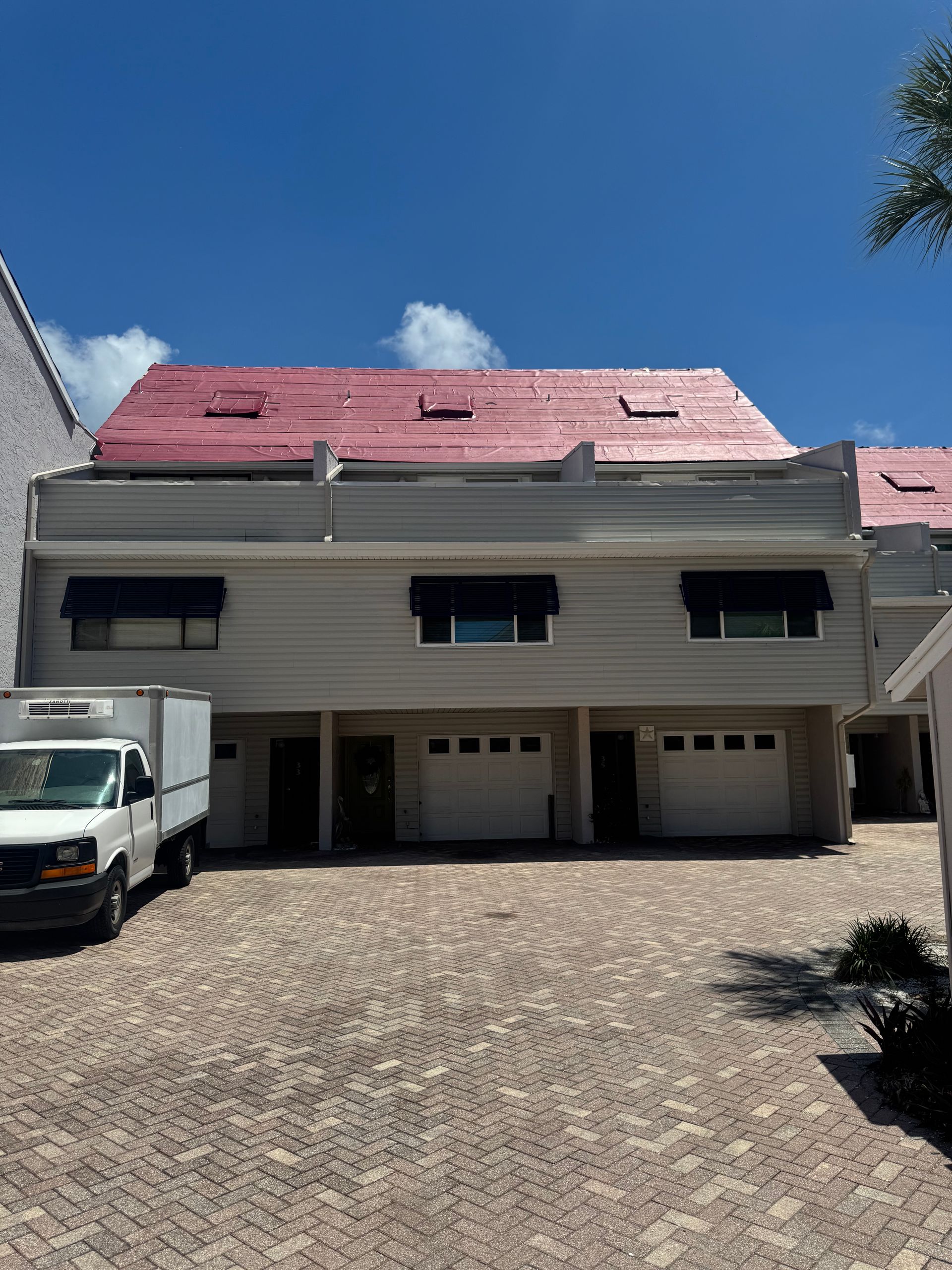 Townhouses with pinkish-red roof, tan siding, black awnings over windows, and white garage doors on a brick driveway.