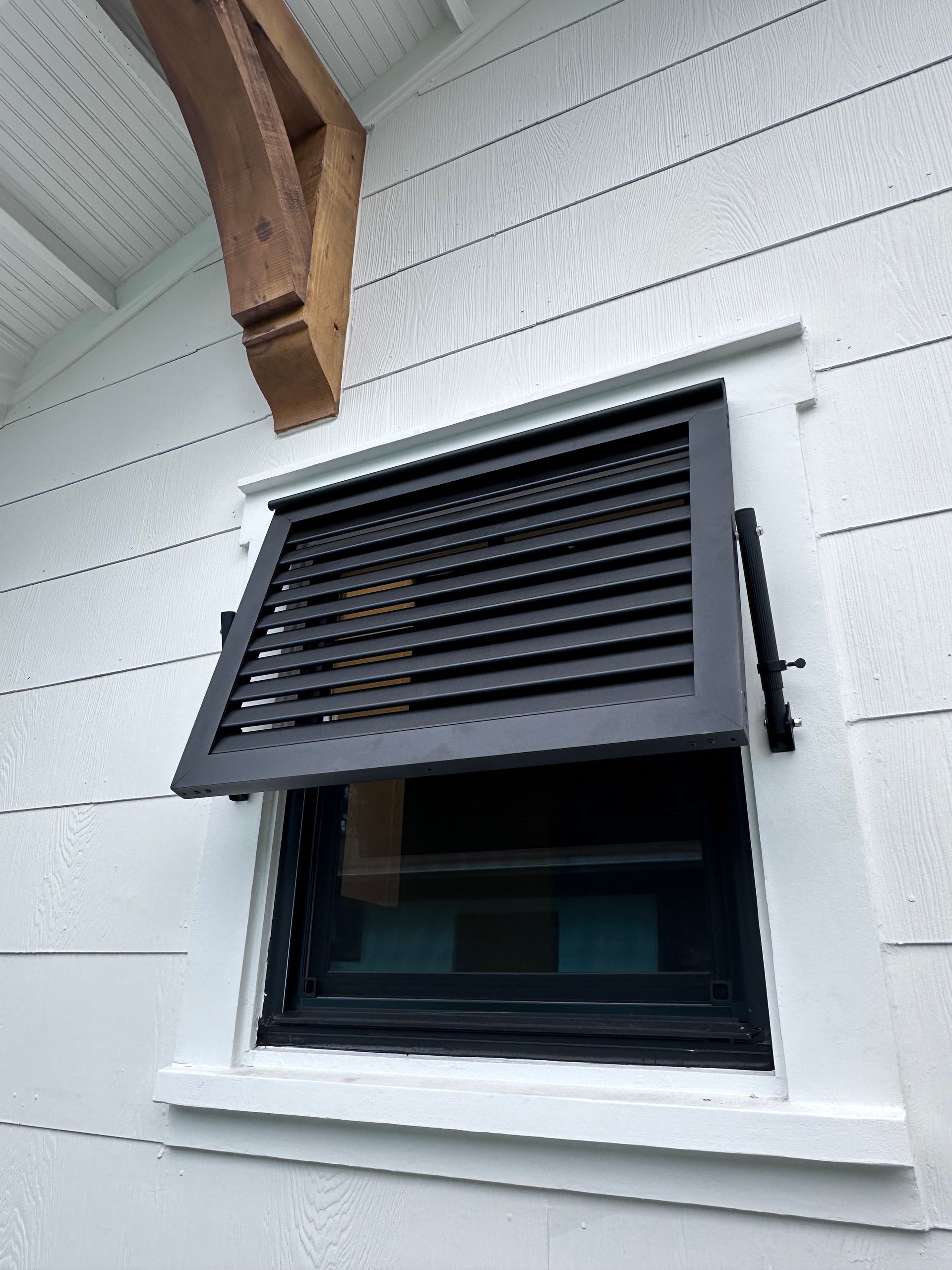 Black louvered window shutter open on a white house with a brown beam.