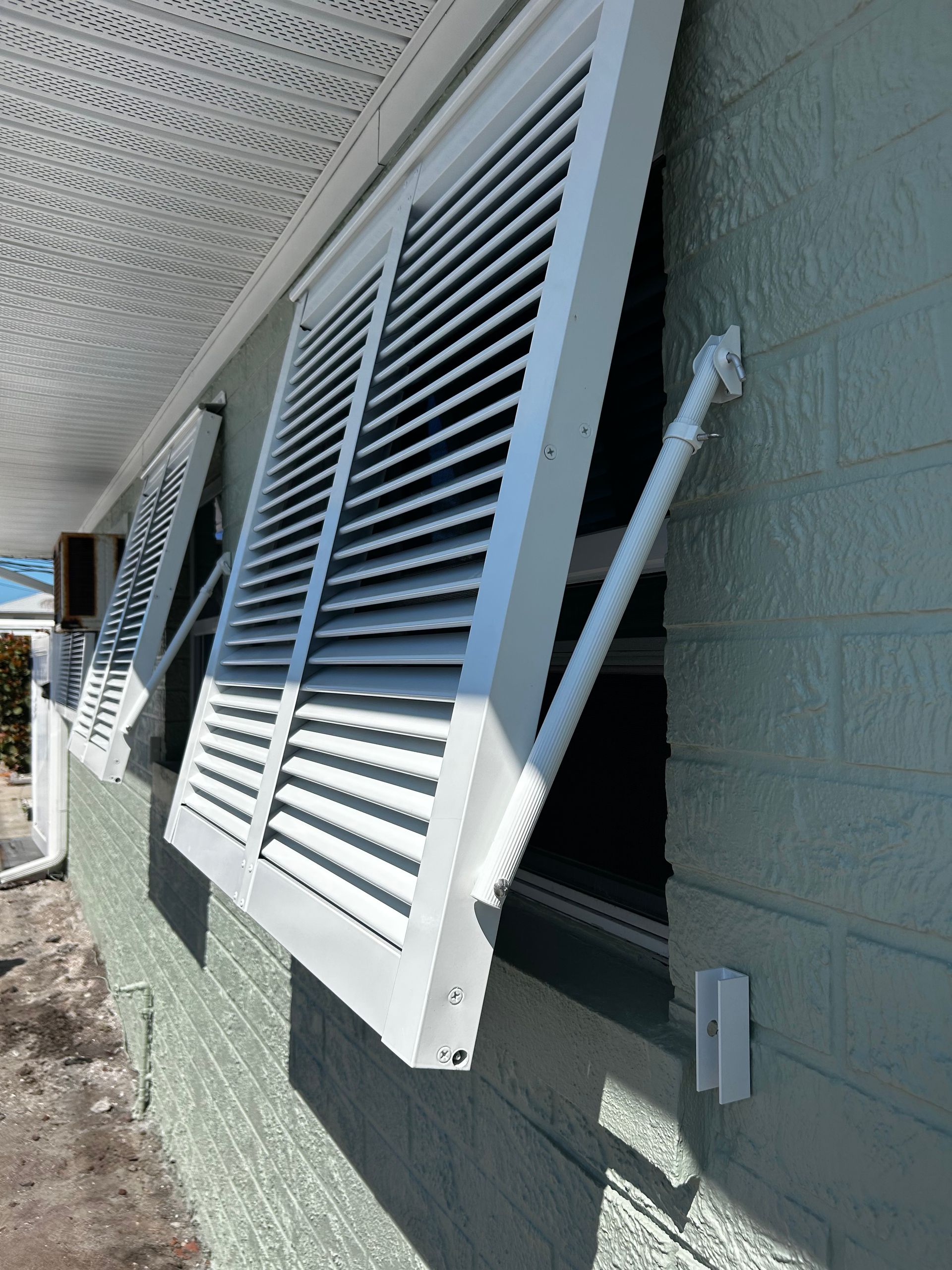 White louvered shutters on a green brick building are open, held by support arms.