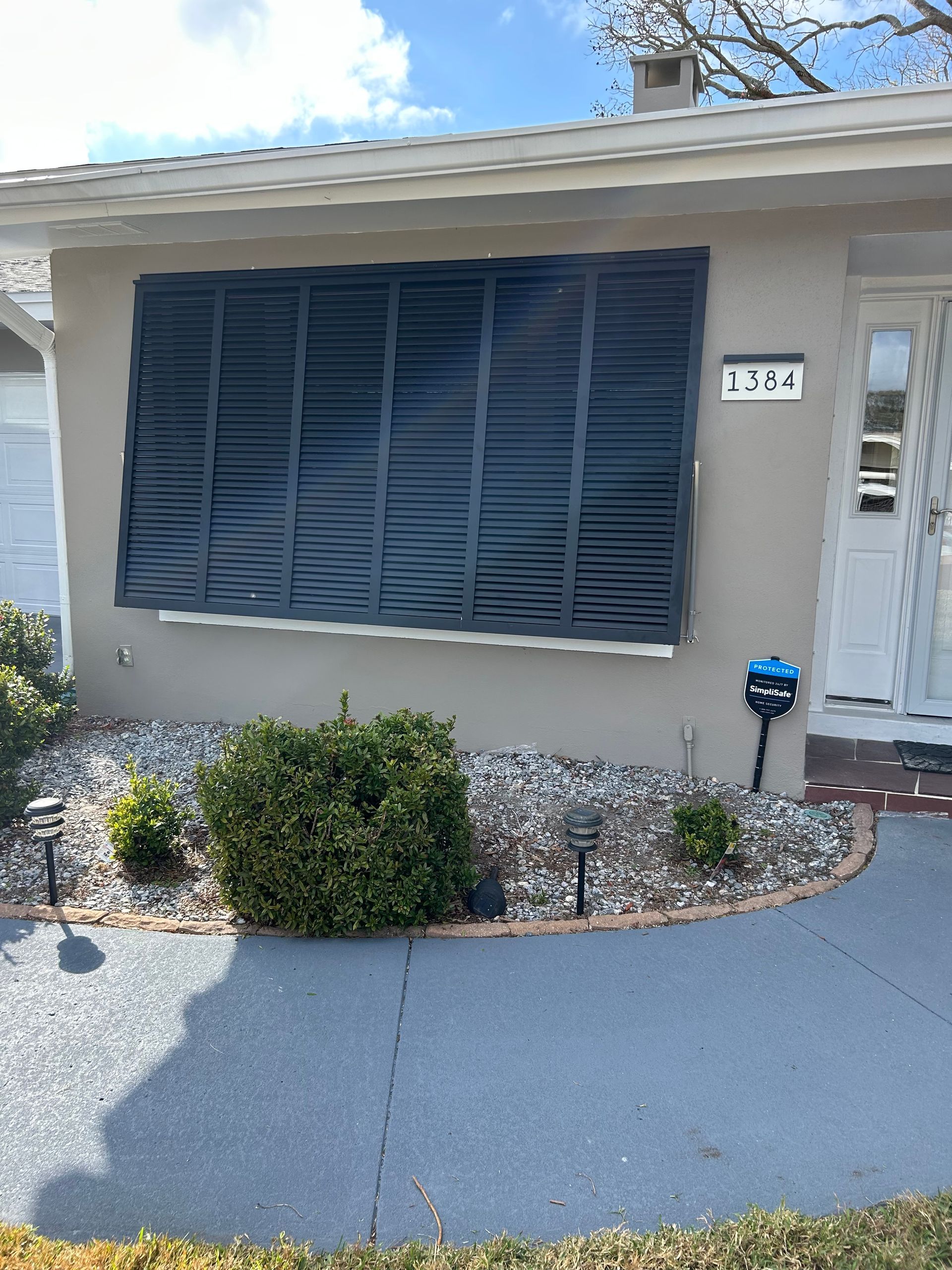 Tan house with black shutter-covered window, small shrubs, gray walkway.