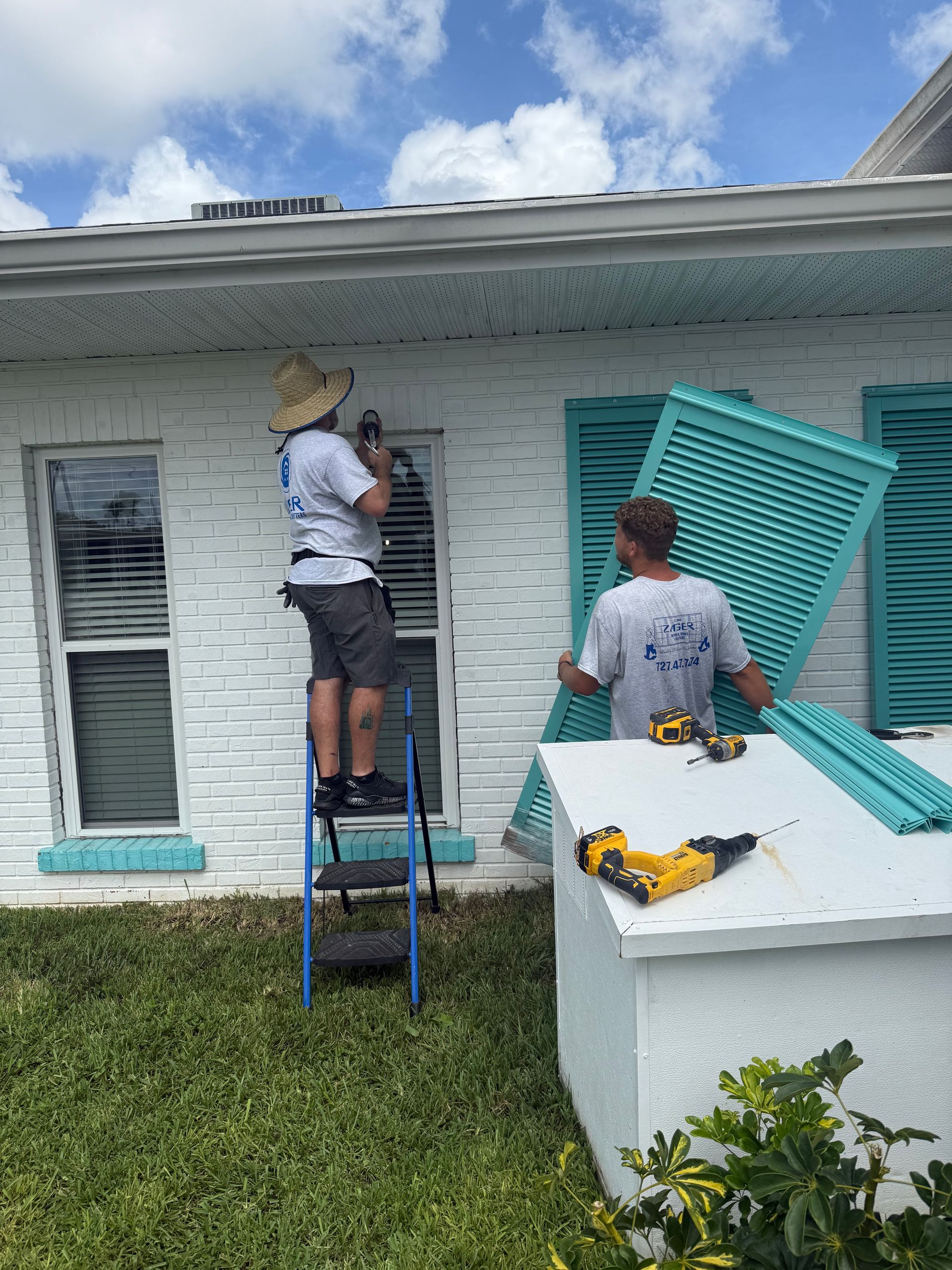 Two people installing teal shutters on a white brick building. One on a ladder, the other holding a shutter.