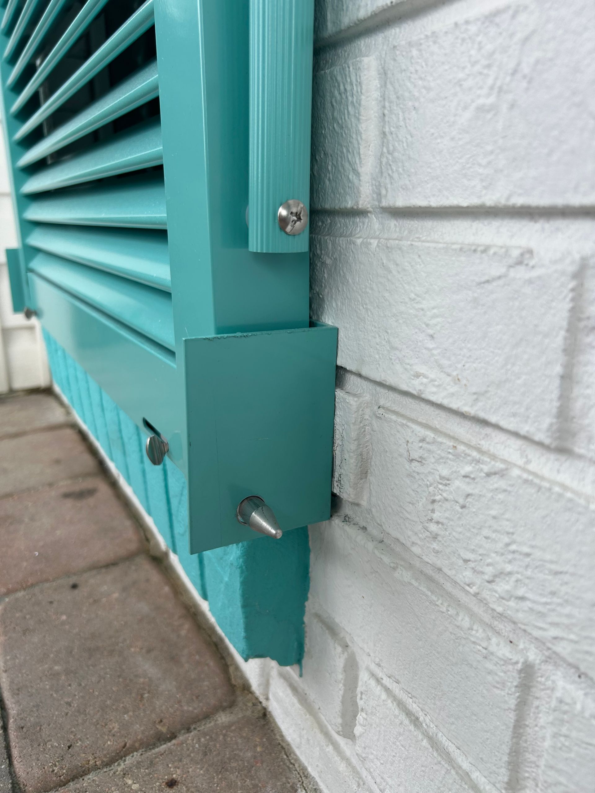 Teal shutter attached to a white brick wall with metal fasteners.