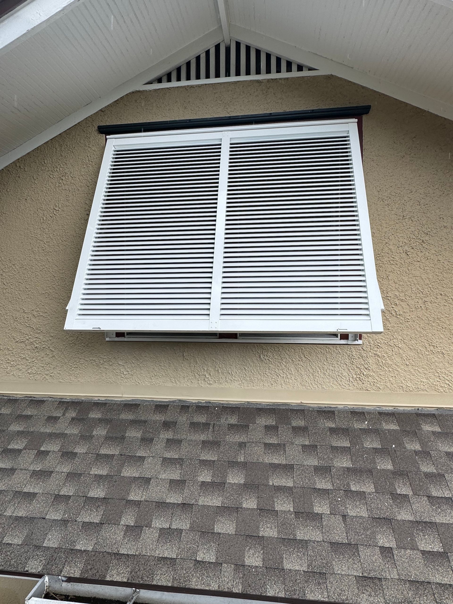 White louvered window with black tubing on tan stucco wall above a shingled roof.