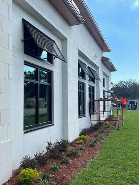 White building with black-framed windows and awnings, green grass, and small plants.