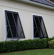 Two windows with black, open shutters on a beige house, next to a green hedge.