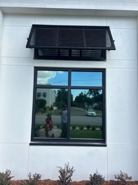 Window with black awning, reflecting sky and people, set in a white building.