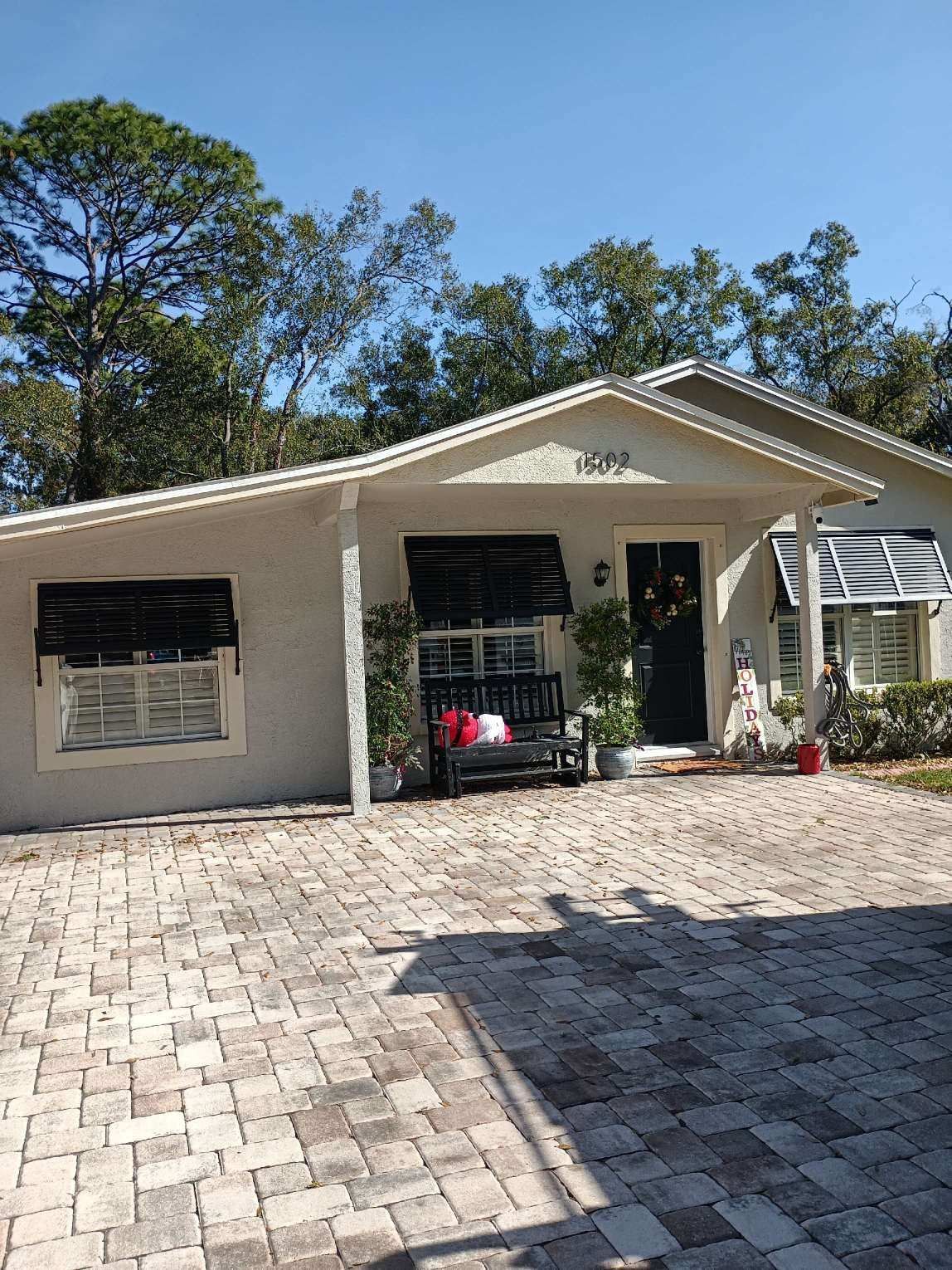 Single-story house with a brick driveway, porch swing, and black window awnings.