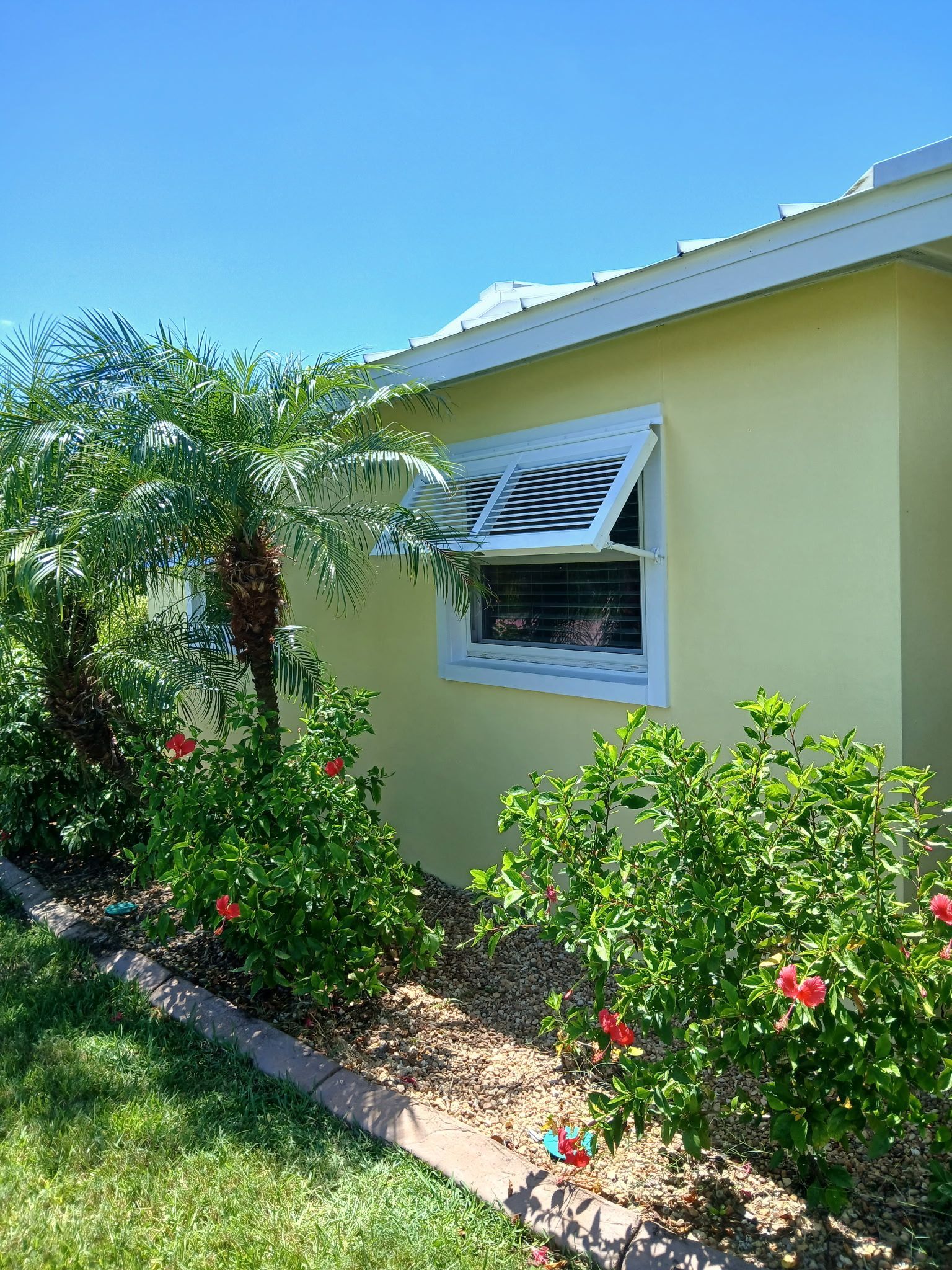 Yellow house with white window shutters open; red flowers in garden bed.