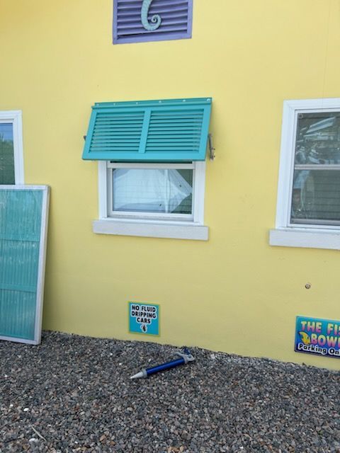 Yellow building with turquoise shutters, one open above a window. A blue pen lies on the gravel ground.