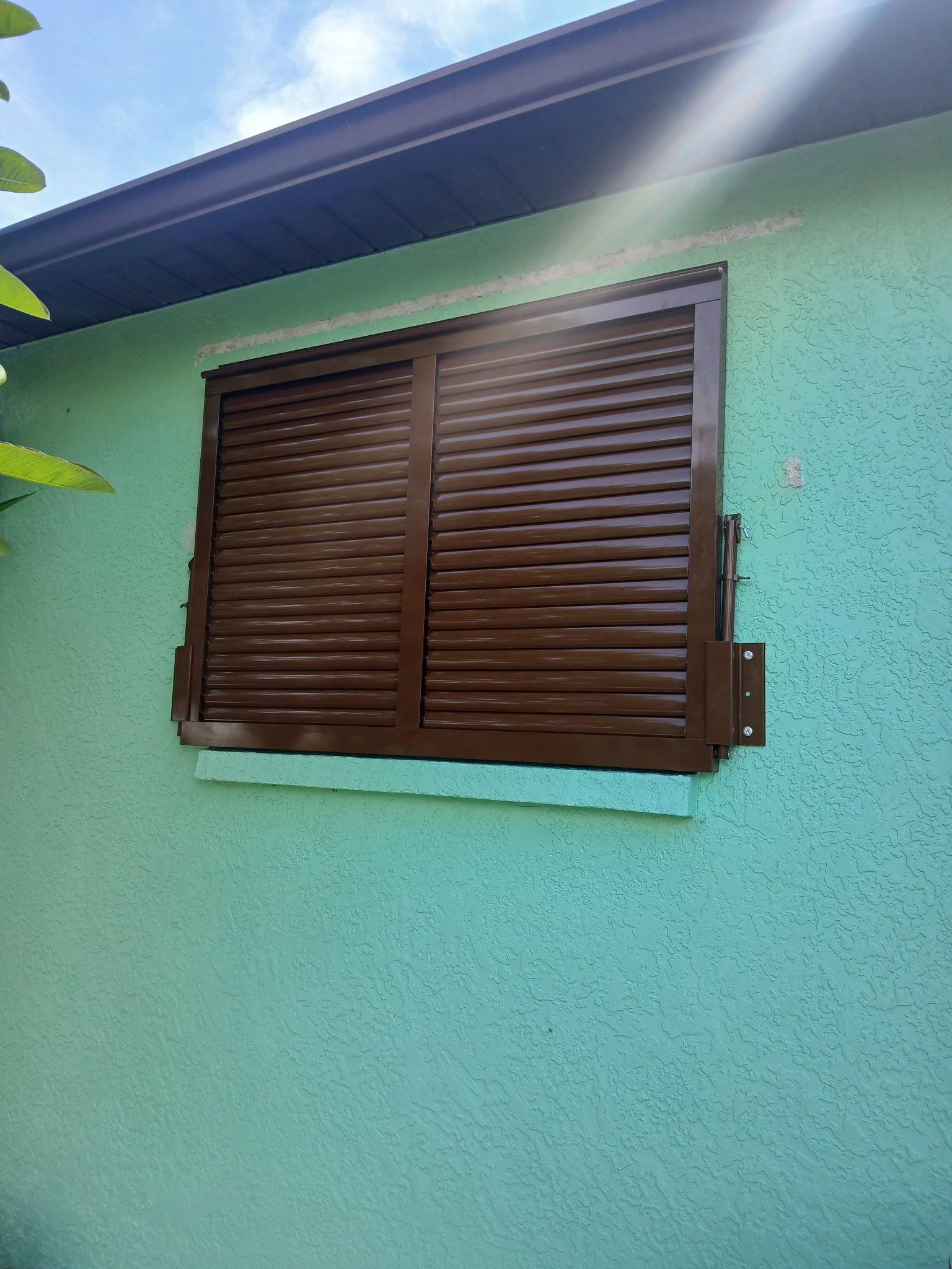 Brown shuttered window on a turquoise stucco wall, under a dark roof.