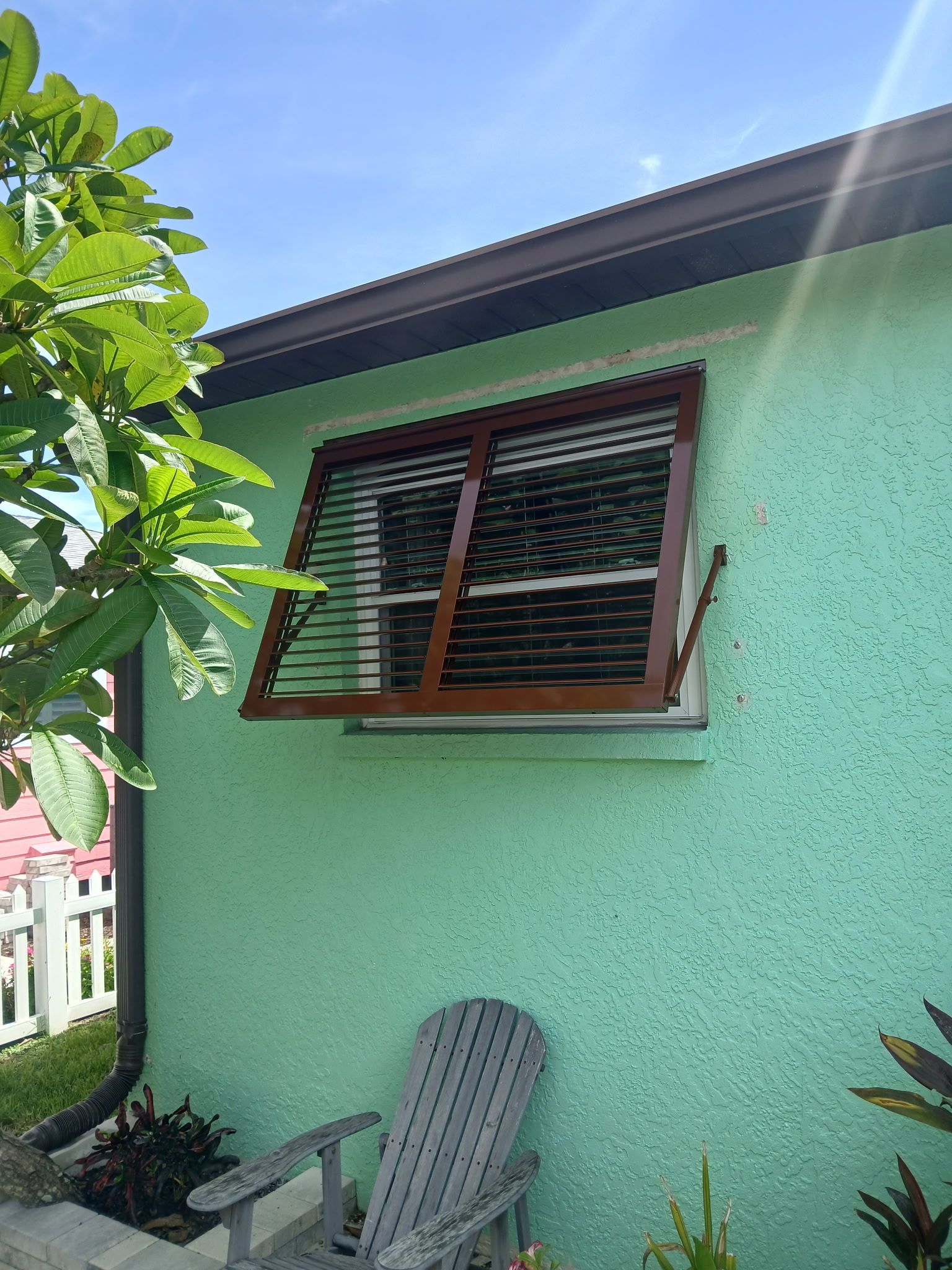 Brown louvered shutters over a window on a light green building. A wooden chair sits below the window.