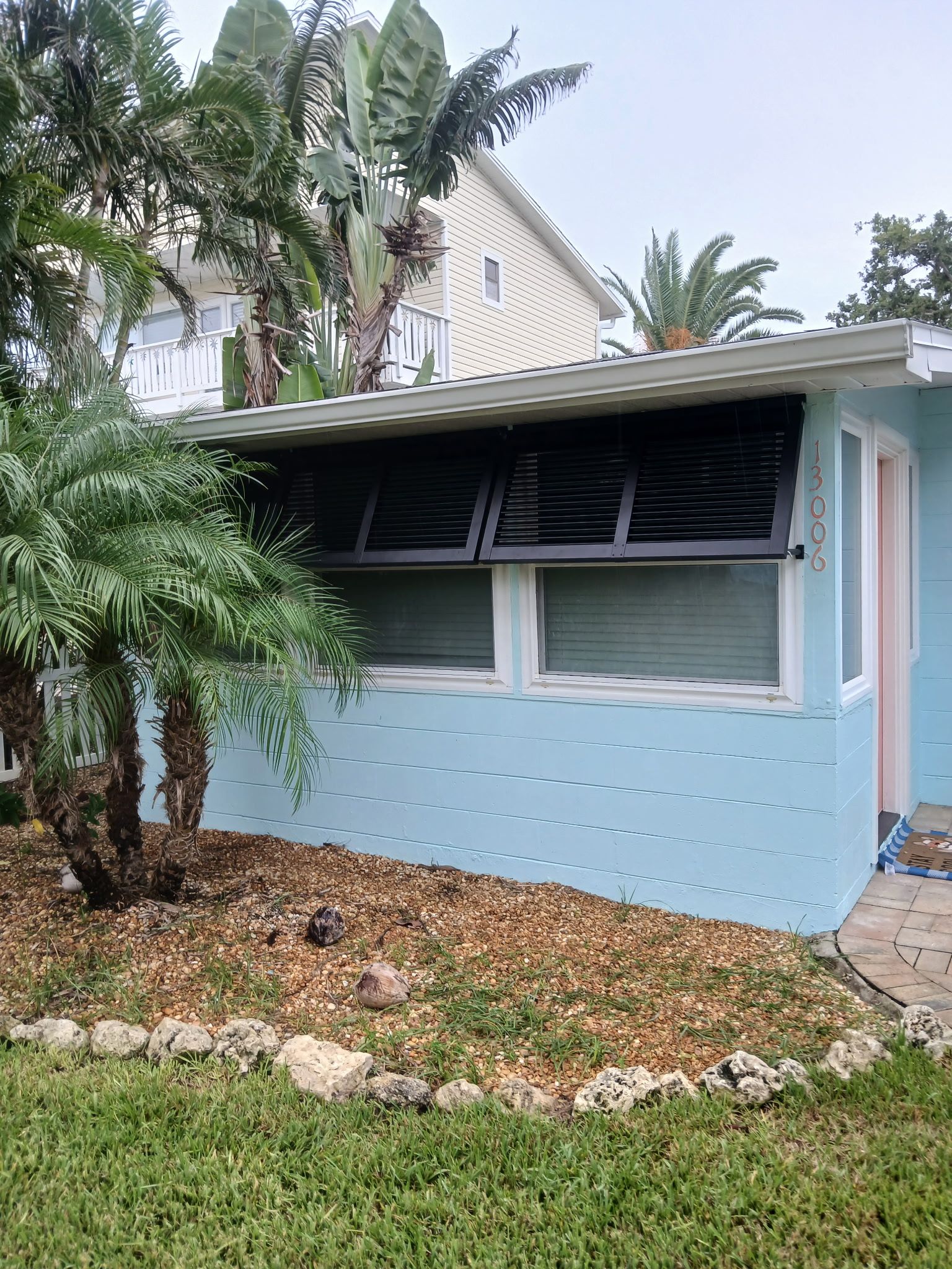 Blue house with black awnings, windows, and palm trees. Brown mulch and grass in front.