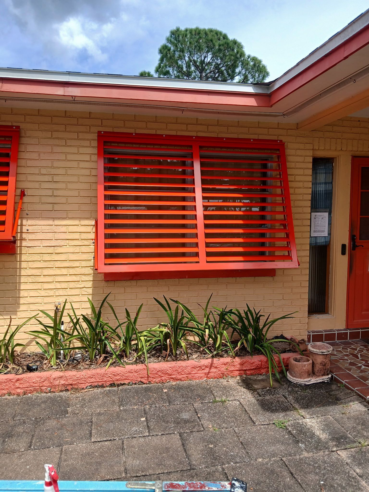 Orange shutters on a yellow brick building, a small garden with plants, and a cloudy sky.
