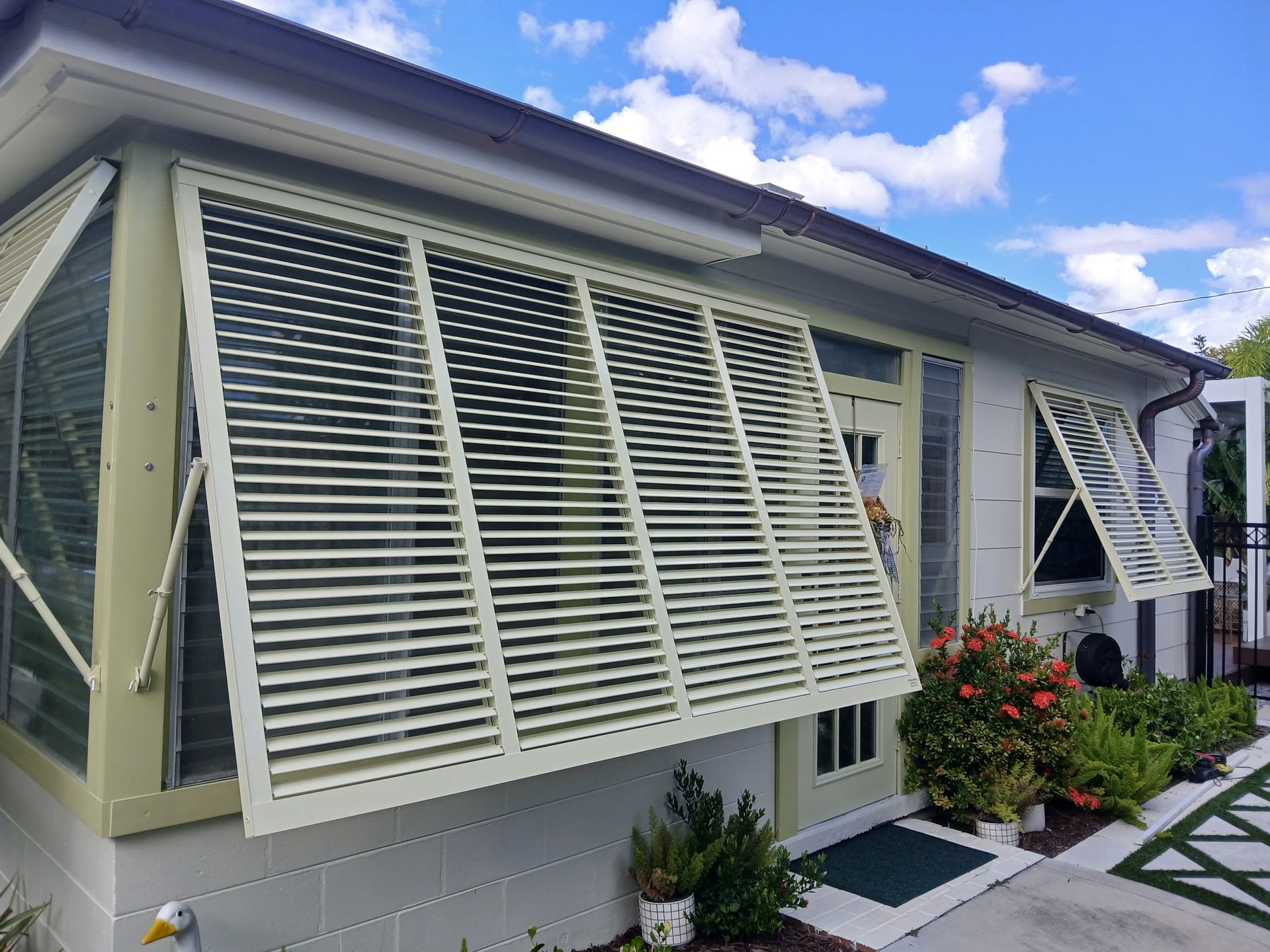 Beige shutters on a white house, partially open. Blue sky, greenery, and a sidewalk visible.