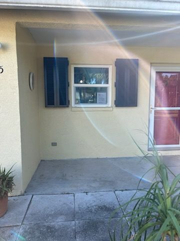 Tan building with window, black shutters, and red door. Potted plants on the porch.