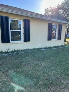 Tan house with black shutters, two windows, and brown roof on a sunny day. Green grass in front.