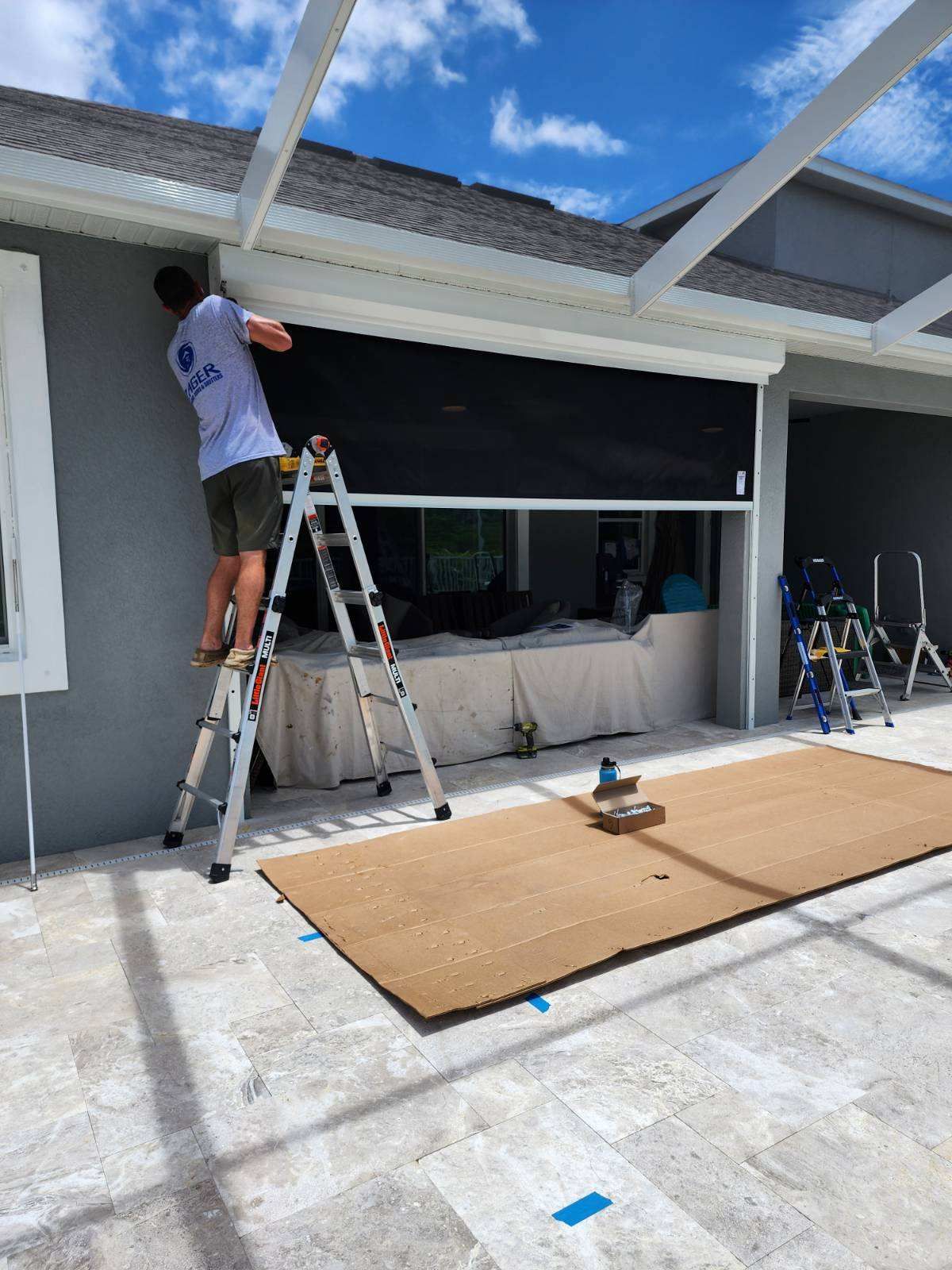 Man on ladder installing a black roll-down shutter on a house exterior under a blue sky.