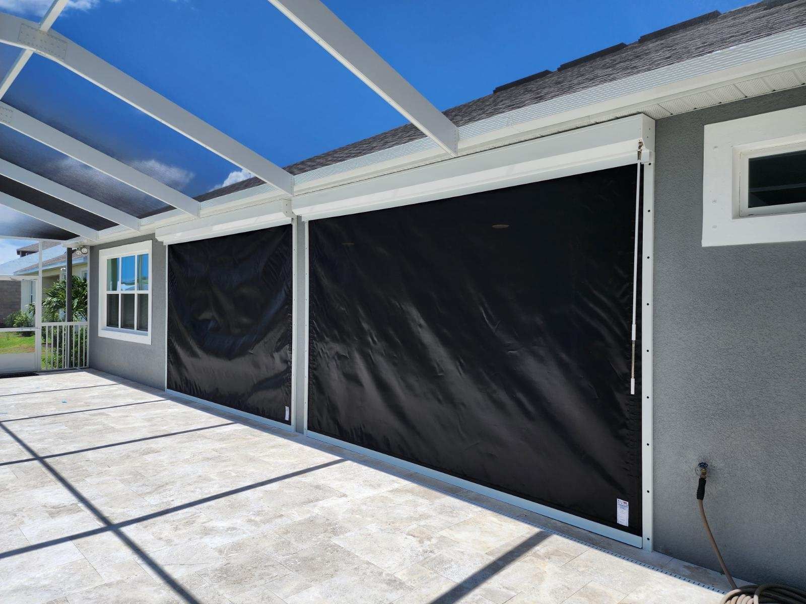 Black roll-down shutters on an outdoor patio, under a white pergola, against a stucco wall.