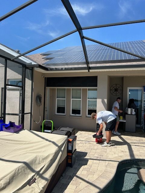 Two men working on a patio, under a screened enclosure. Solar panels on roof. Patio furniture covered. Sunny day.