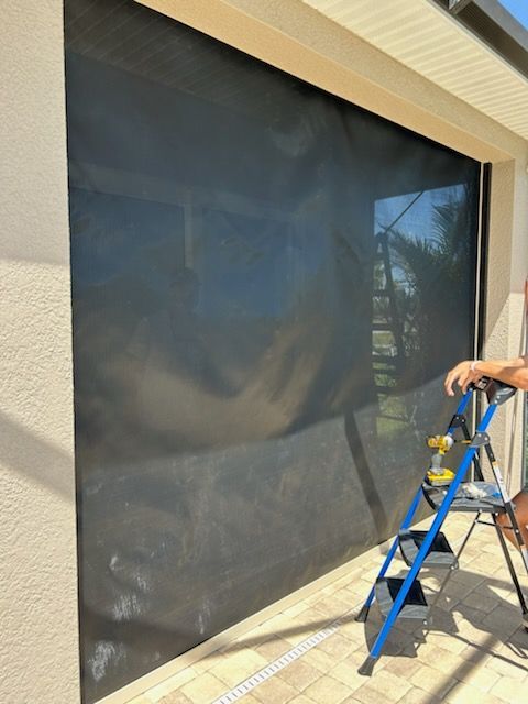 A person on a small ladder installing a large black screen over a window on a stucco wall.